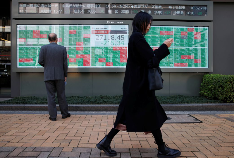 A woman walks past a man examining an electronic board showing Japan's Nikkei average and stock quotations outside a brokerage, in Tokyo, Japan, March 20, 2023. REUTERS/Androniki Christodoulou/File Photo